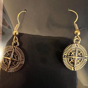 Puede incluir: Un par de pendientes dorados con un diseño de rosa de los vientos. Los pendientes cuelgan de ganchos dorados y se muestran sobre un fondo negro. El texto "Handmade with love" es visible en la parte superior.