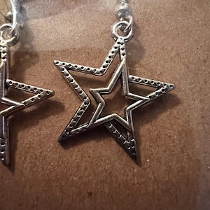 May include: Silver-toned star-shaped earrings. Each earring features a layered star design with a dotted border and a fish hook closure. The earrings are displayed against a brown background.