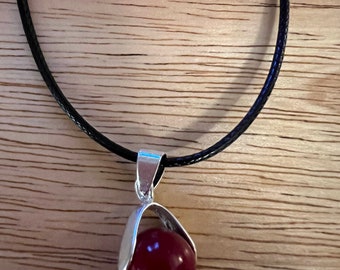 May include: A silver pendant necklace with a dark red gemstone set in a silver cage. The necklace is on a black cord.