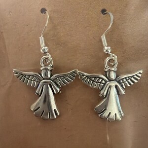 May include: A pair of silver-colored angel earrings. Each earring features a detailed angel figure with outstretched wings and a flowing gown. The earrings have a simple hook closure.