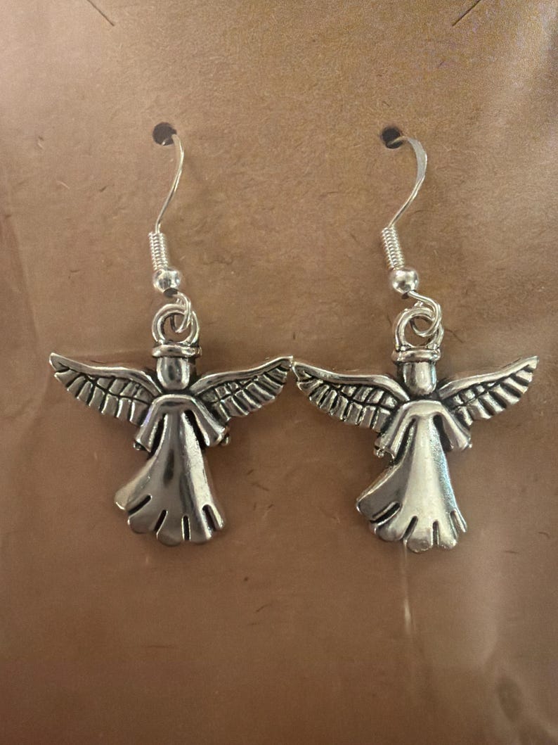 May include: A pair of silver-colored angel earrings. Each earring features a detailed angel figure with outstretched wings and a flowing gown. The earrings have a simple hook closure.