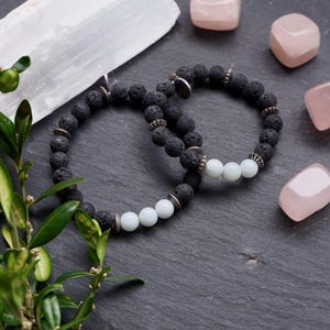 May include: Two black lava stone bracelets with white beads. The bracelets are on a dark gray surface with pink and white stones.