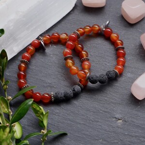 May include: Two bracelets with orange carnelian beads and black lava rock beads. The bracelets are on a dark grey surface.