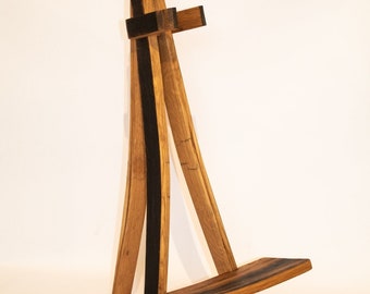 May include: A wooden easel made from repurposed wine barrel staves. The easel has a dark brown stain and a natural wood finish. It is a triangular shape with a flat top shelf.
