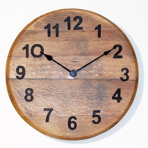 Puede incluir: Un reloj de madera con números y manecillas negros. La esfera del reloj está hecha de madera reciclada con un acabado rústico.