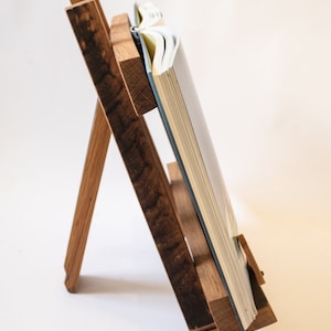 May include: A wooden book stand with a book resting on it. The stand is made of dark wood with a natural finish. The book is open and the pages are visible.