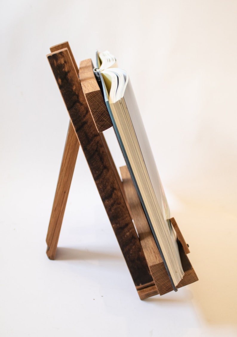 May include: A wooden book stand with a book resting on it. The stand is made of dark wood with a natural finish. The book is open and the pages are visible.