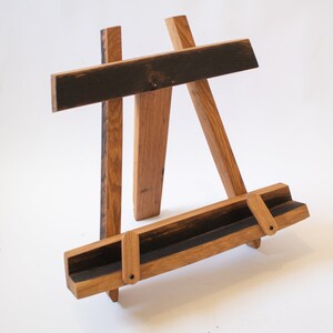 May include: A wooden book stand made from reclaimed whiskey barrel staves. The stand has a dark brown finish and features a unique design with a curved base and a flat top.