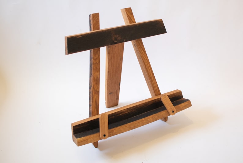 May include: A wooden book stand made from reclaimed whiskey barrel staves. The stand has a dark brown finish and features a unique design with a curved base and a flat top.