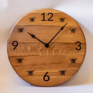 Puede incluir: Un reloj de madera redondo con manecillas negras y diseños de abejas. La esfera del reloj tiene números negros 12, 9, 6 y 3.