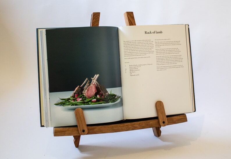 May include: A wooden cookbook stand with a cookbook open to a recipe for rack of lamb. The recipe includes a list of ingredients and instructions. The cookbook is propped open on the stand, which has two wooden arms that hold the book in place.