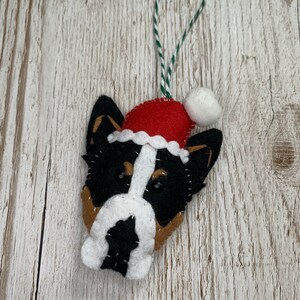 May include: A felt Christmas ornament shaped like a dog's head wearing a red Santa hat with a white pom-pom. The dog has black, white, and brown fur.