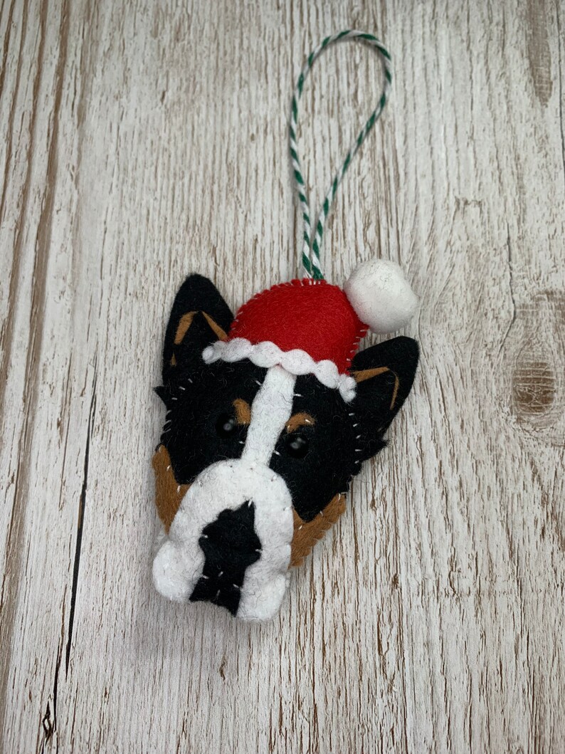 May include: A felt Christmas ornament shaped like a dog's head wearing a red Santa hat with a white pom-pom. The dog has black, white, and brown fur.
