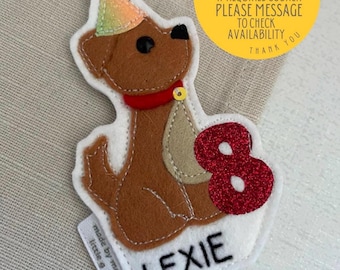 May include: A brown felt dog wearing a party hat and holding a red felt number 8. The dog is sitting on a white felt background with the name "Lexie" embroidered below. The text "made by me little a" is visible in the lower left corner. A yellow circle with the text "2 week making time if required sooner please message to check availability thank you" is in the upper right corner.