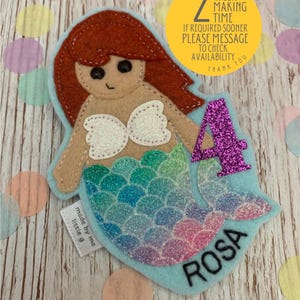 May include: A felt mermaid appliqué with a light blue tail, pink and blue glitter scales, and a white seashell bra. The mermaid has red hair and brown eyes. The appliqué is on a light blue background with the number 4 in purple glitter and the name "ROSA" embroidered in black. The text "2 WEEK MAKING TIME IF REQUIRED SOONER PLEASE MESSAGE TO CHECK AVAILABILITY THANK YOU" is on a yellow circle.