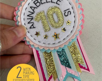 May include: A white felt circle with pink and blue scalloped edges. The circle has a gold glitter number 10 and the text "Annabelle is" in black. The circle is attached to a pink, blue, and gold glitter ribbon.