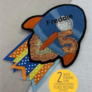 May include: A blue and orange felt rocket with the name "Freddie" and the number "5" on it. The rocket is attached to a string of blue, orange and yellow ribbon with white polka dots. A yellow circle with the text "2 week making time if required sooner please message to check availability thank you" is in the lower right corner of the image.
