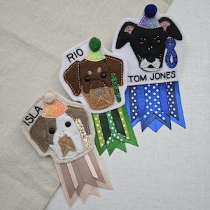 May include: Three felt dog-shaped badges with party hats and ribbons. Each badge features a different dog breed and a name: Isla, Rio, and Tom Jones. The ribbons are in various colors, including blue, green, and tan.
