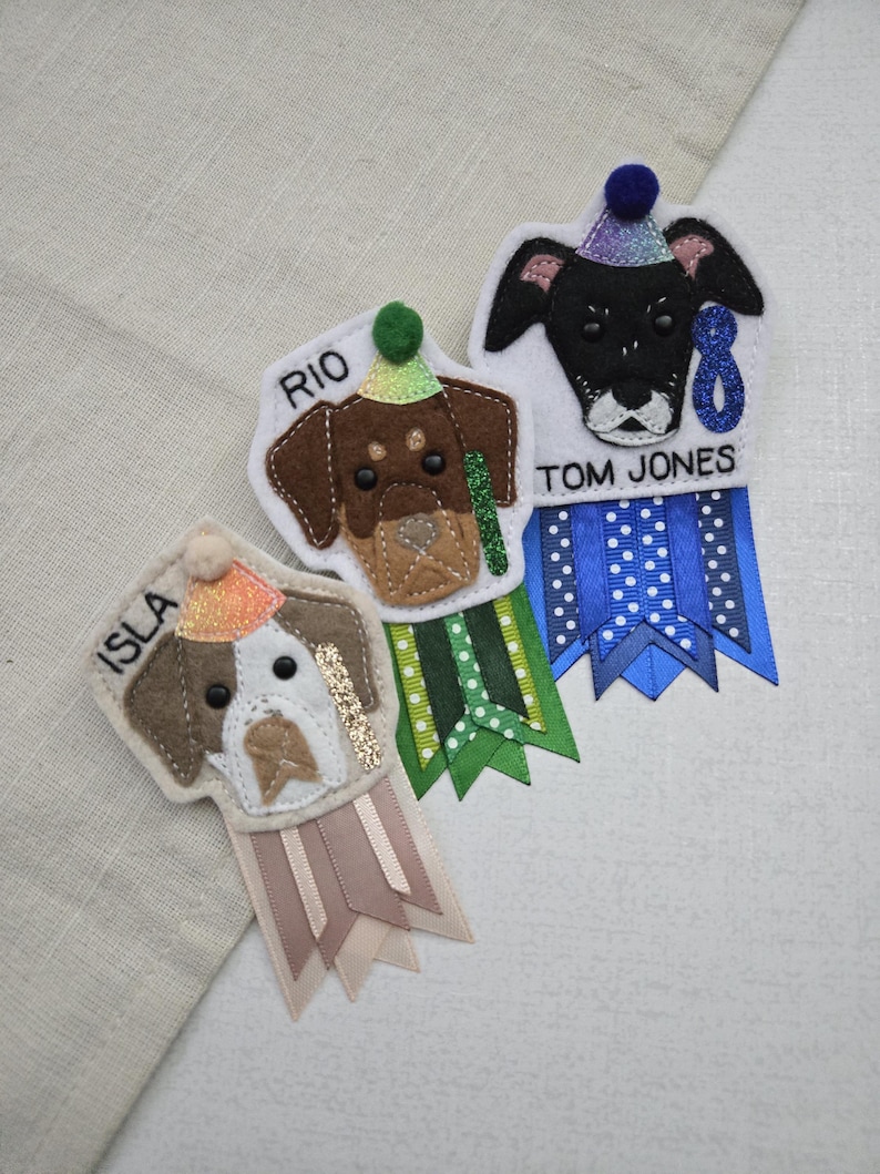 May include: Three felt dog-shaped badges with party hats and ribbons. Each badge features a different dog breed and a name: Isla, Rio, and Tom Jones. The ribbons are in various colors, including blue, green, and tan.