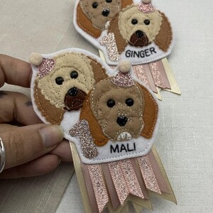 May include: Handmade felt dog-themed birthday badges. Each badge features a dog's face, a glittery number 1, and a name tag. The badges have pink and cream ribbons. One badge says "Ginger" and the other says "Mali".