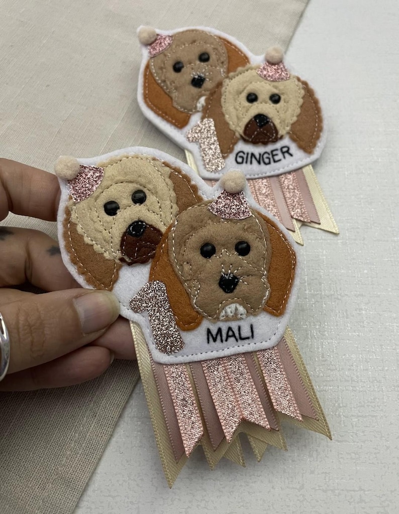 May include: Handmade felt dog-themed birthday badges. Each badge features a dog's face, a glittery number 1, and a name tag. The badges have pink and cream ribbons. One badge says "Ginger" and the other says "Mali".