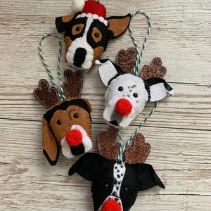 May include: Four felt Christmas ornaments shaped like dogs with reindeer antlers and Santa hats. The ornaments are brown, black, and white with red noses.