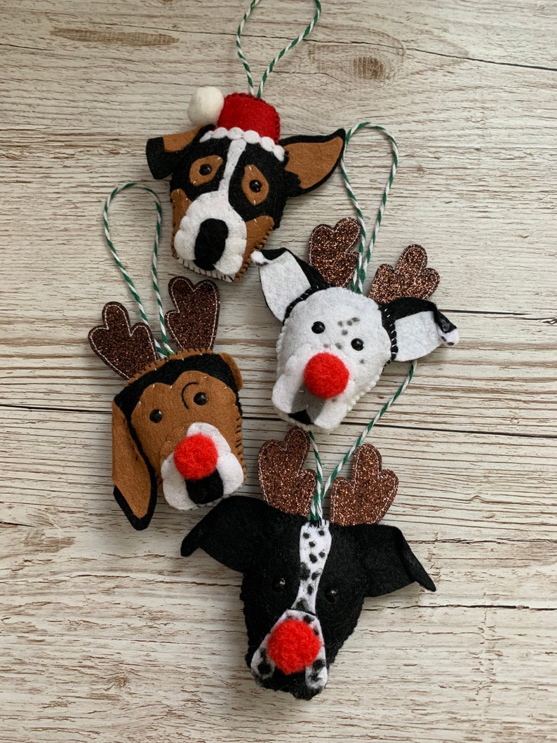 May include: Four felt Christmas ornaments shaped like dogs with reindeer antlers and Santa hats. The ornaments are brown, black, and white with red noses.