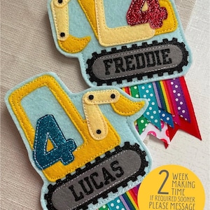 May include: Two felt appliqué birthday decorations shaped like yellow construction vehicles with the numbers 4 and the names Lucas and Freddie. The decorations are attached to colourful ribbon.
