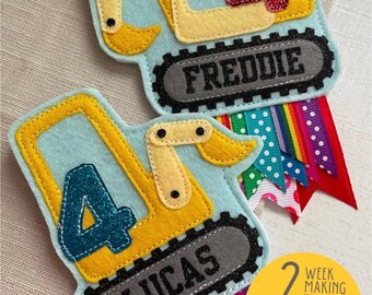 May include: Two felt appliqué birthday decorations shaped like yellow construction vehicles with the numbers 4 and the names Lucas and Freddie. The decorations are attached to colorful ribbon.