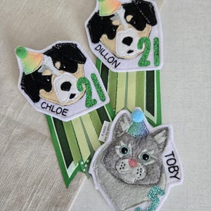 May include: Three felt pet birthday badges. Two feature black and white dogs with party hats and the number 21, with the names "Chloe" and "Dillon". The third badge is a gray cat with a blue party hat and the name "Toby" and the number 3.