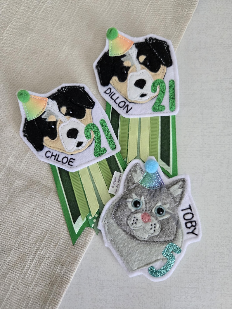 May include: Three felt pet birthday badges. Two feature black and white dogs with party hats and the number 21, with the names "Chloe" and "Dillon". The third badge is a gray cat with a blue party hat and the name "Toby" and the number 3.