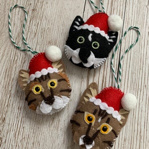 May include: Three felt cat ornaments with red Santa hats and white pom-poms. The cats have different colored fur: black, brown, and tabby.