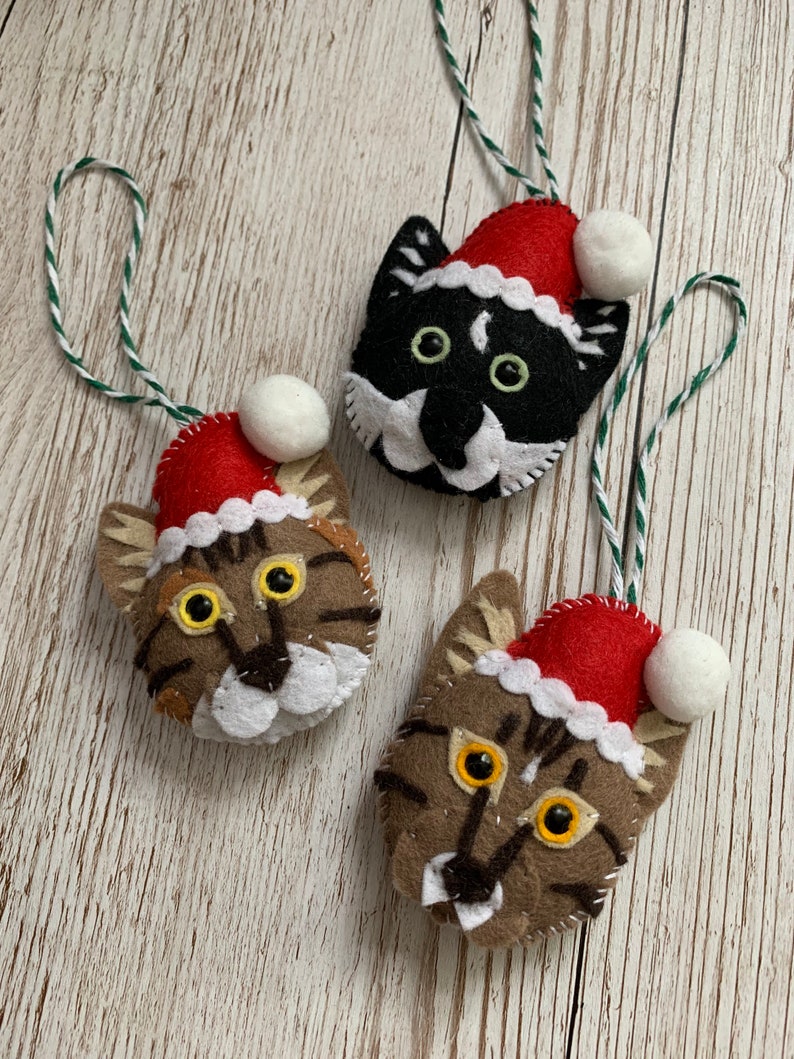 May include: Three felt cat ornaments with red Santa hats and white pom-poms. The cats have different colored fur: black, brown, and tabby.