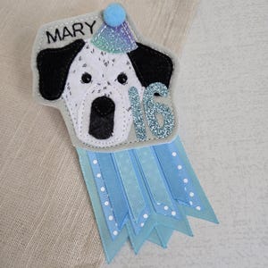 May include: A handmade felt birthday badge featuring a dalmatian dog design wearing a party hat. The badge has the name "MARY" and the number "16" in glitter. Light blue ribbons with white polka dots and solid light blue ribbons hang below the dog.