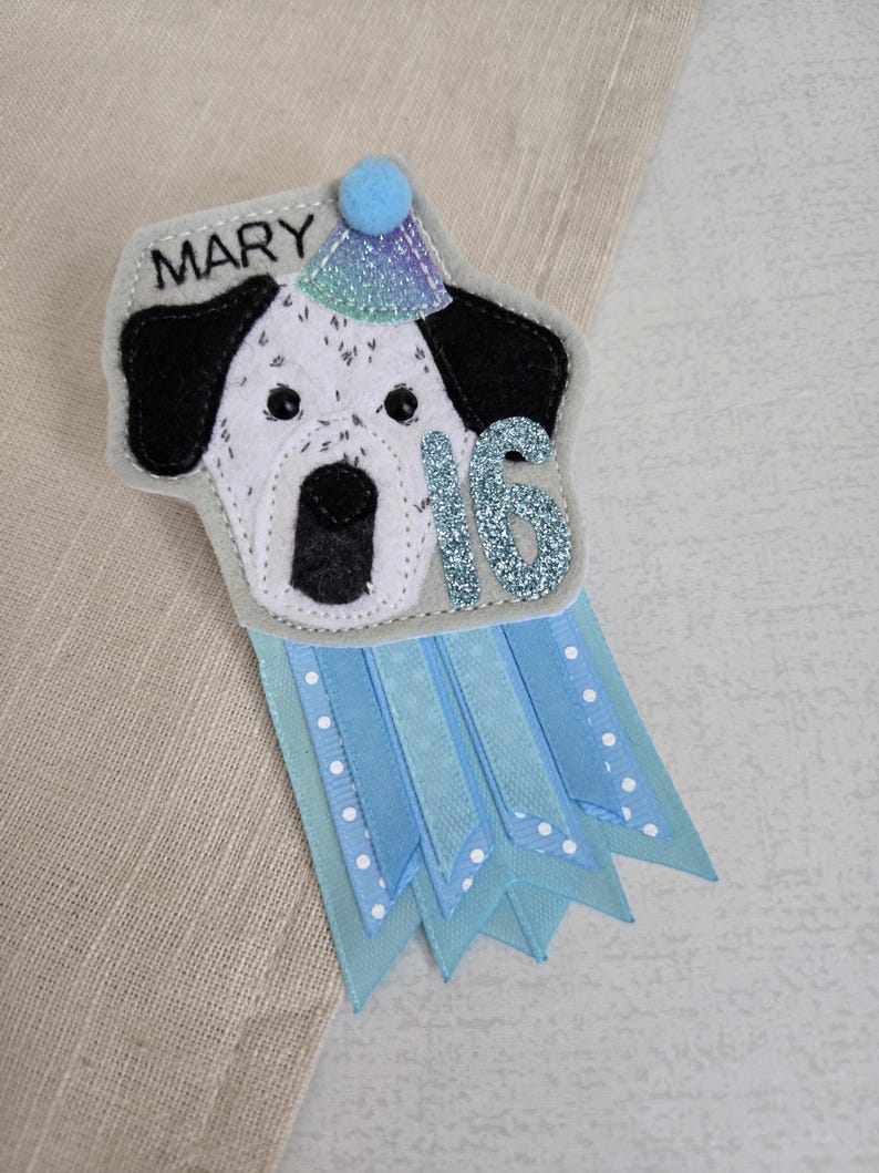May include: A handmade felt birthday badge featuring a dalmatian dog design wearing a party hat. The badge has the name "MARY" and the number "16" in glitter. Light blue ribbons with white polka dots and solid light blue ribbons hang below the dog.