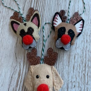 May include: Three felt Christmas ornaments shaped like dogs with reindeer antlers. The dogs are beige with black eyes and red noses. The antlers are brown with glitter.