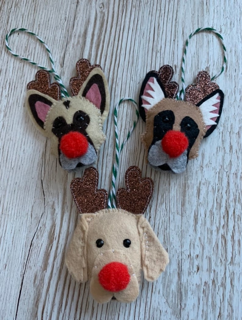 May include: Three felt Christmas ornaments shaped like dogs with reindeer antlers. The dogs are beige with black eyes and red noses. The antlers are brown with glitter.