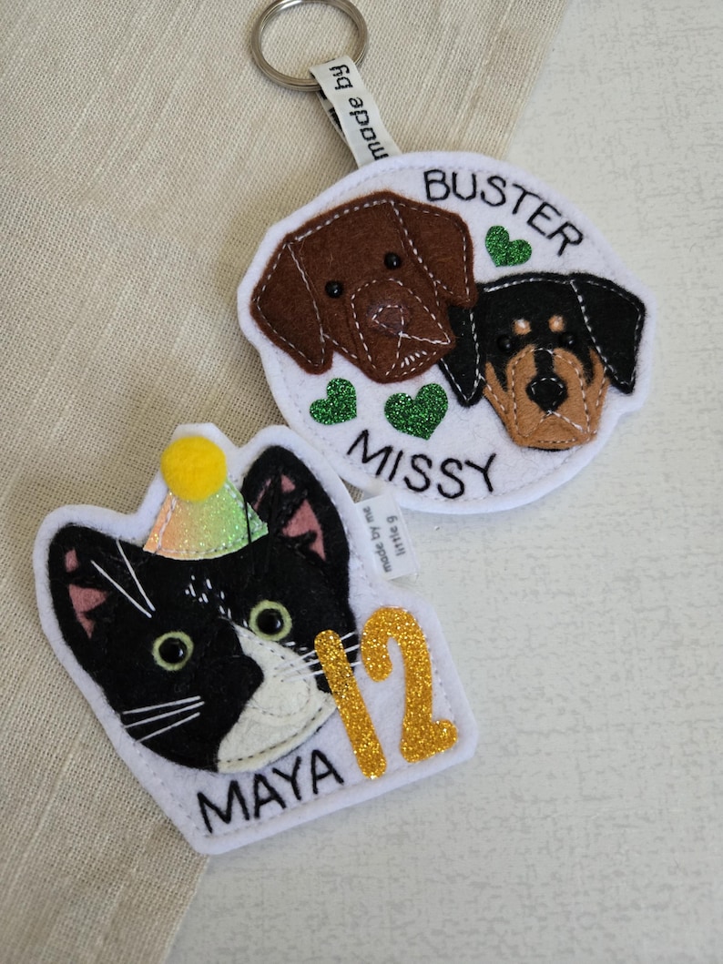 May include: Two felt keychains: one with a brown and black dog design, the names "Buster" and "Missy", and green heart accents. The other features a black and white cat with a party hat, the name "Maya", and the number "12" in gold glitter.