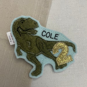 May include: A light blue felt patch with a green felt dinosaur, the name "COLE" in black embroidery, and a gold glitter number "2". The patch is made by "The Little B".
