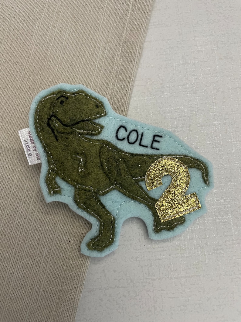 May include: A light blue felt patch with a green felt dinosaur, the name "COLE" in black embroidery, and a gold glitter number "2". The patch is made by "The Little B".