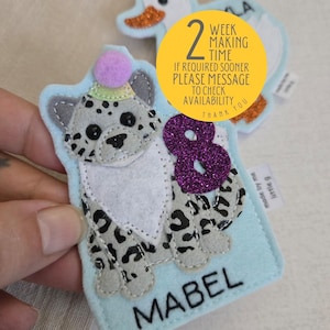 May include: A light blue felt patch featuring a grey leopard with black spots, a purple glitter number eight, and a purple pom-pom on its head. The name MABEL is embroidered in black at the bottom. A yellow circular sticker displays 2 WEEK MAKING TIME.