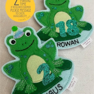 May include: Two felt frog appliques with glitter numbers, one with the number 2 and the name Angus, the other with the number 18 and the name Rowan. Both frogs are wearing party hats and are on a light blue background.