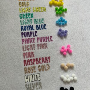 May include: A white surface displays a color chart with glittery text labels and small pom-poms. Colors include red, orange, gold, green, blue, purple, pink, raspberry, rose gold, white, silver, and black. The pom-poms match the color labels.