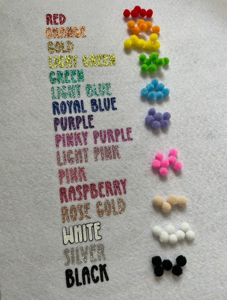 May include: A white surface displays a color chart with glittery text labels and small pom-poms. Colors include red, orange, gold, green, blue, purple, pink, raspberry, rose gold, white, silver, and black. The pom-poms match the color labels.