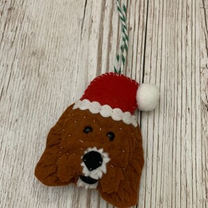 May include: A felt Christmas ornament shaped like a dog wearing a red Santa hat with a white pom-pom. The dog has brown fur, black eyes, and a black nose.
