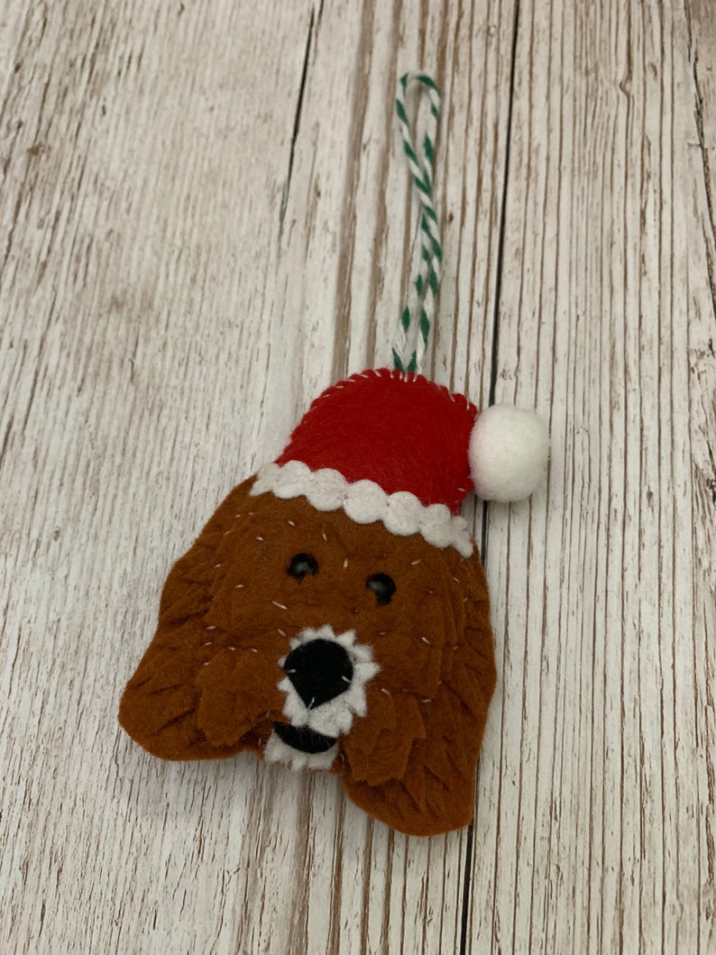 May include: A felt Christmas ornament shaped like a dog wearing a red Santa hat with a white pom-pom. The dog has brown fur, black eyes, and a black nose.