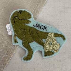 May include: A light blue felt dinosaur applique with a green dinosaur, the name "JACK" and the number "4" in gold glitter.