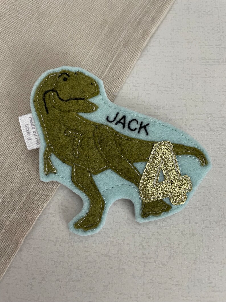 May include: A light blue felt dinosaur applique with a green dinosaur, the name "JACK" and the number "4" in gold glitter.
