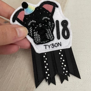 May include: A handmade felt birthday badge featuring a black dog illustration wearing a party hat. The badge includes the name "TYSON" and the number "18" in glitter. Black and white ribbons with polka dots and a scalloped edge complete the design.