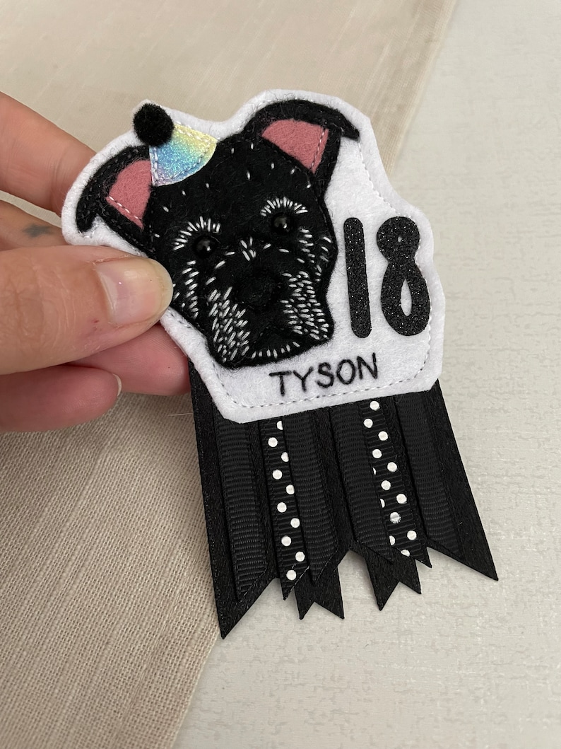 May include: A handmade felt birthday badge featuring a black dog illustration wearing a party hat. The badge includes the name "TYSON" and the number "18" in glitter. Black and white ribbons with polka dots and a scalloped edge complete the design.